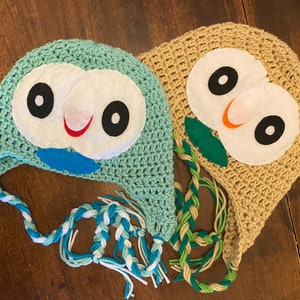 May include: Two crocheted owl hats. One is light blue with white, black, red, and blue felt details. The other is tan with white, black, and orange felt details. Both have braided ties.
