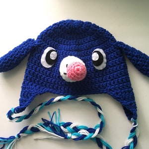 May include: A handmade blue crochet hat with a cartoon animal design. The hat features white and black eyes, a pink nose, and blue ears. Braided ties in blue, white, and turquoise extend from the hat.