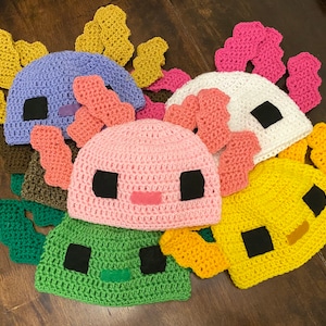 May include: A set of seven crocheted hats featuring axolotl designs. The hats are in various colors, including green, yellow, pink, white, purple, and brown. Each hat has a unique axolotl face with black eyes and a pink mouth.