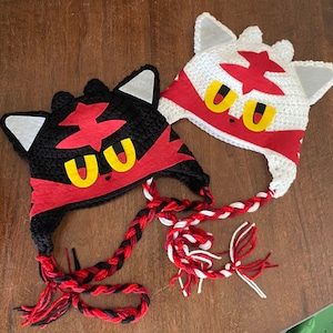 May include: Two crocheted hats with a cat-like design. One hat is black, the other is white. Both have red and yellow accents, with braided ties in red, black, and white. The hats are likely for children or themed costumes.