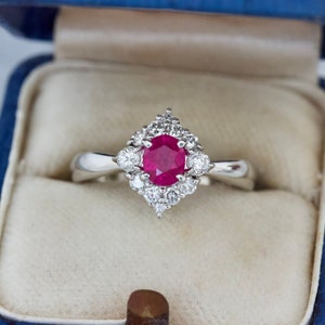 May include: A white gold ring featuring a large, round, red ruby surrounded by smaller diamonds. The ring is displayed in a blue velvet ring box.