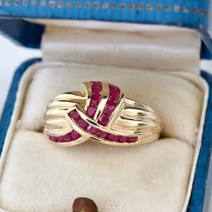 May include: A gold ring with a knot design, set with multiple small, red gemstones. The ring is displayed inside a blue jewelry box with a white interior. The ring's gold band has a ribbed texture.