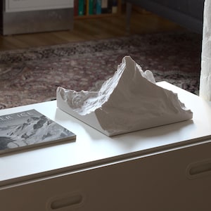 Puede incluir: Una escultura de cer&aacute;mica blanca de una cordillera de monta&ntilde;as sobre una mesa blanca con un libro titulado "Glacier Ice" sobre la mesa.