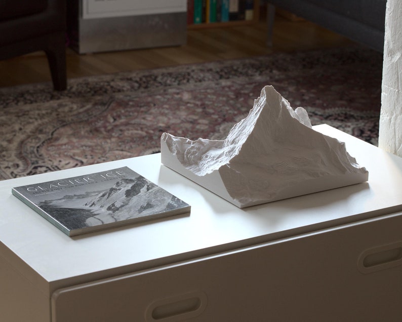 Puede incluir: Una escultura de cer&aacute;mica blanca de una cordillera de monta&ntilde;as sobre una mesa blanca con un libro titulado "Glacier Ice" sobre la mesa.