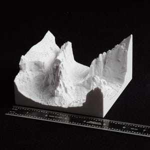 May include: A white, 3D-printed model of a mountain range with detailed textures and sharp peaks. The model is rectangular and sits on a black surface. A ruler is placed next to the model for scale.
