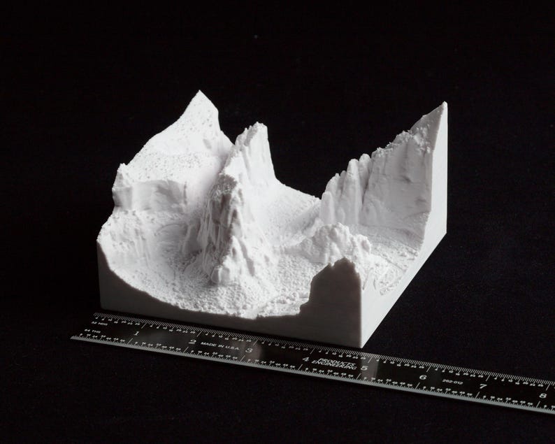May include: A white, 3D-printed model of a mountain range with detailed textures and sharp peaks. The model is rectangular and sits on a black surface. A ruler is placed next to the model for scale.