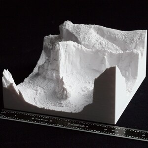 May include: A white 3D-printed model of a mountainous landscape, featuring cliffs, valleys, and textured surfaces. A ruler is placed next to the model for scale. The model is on a black surface.