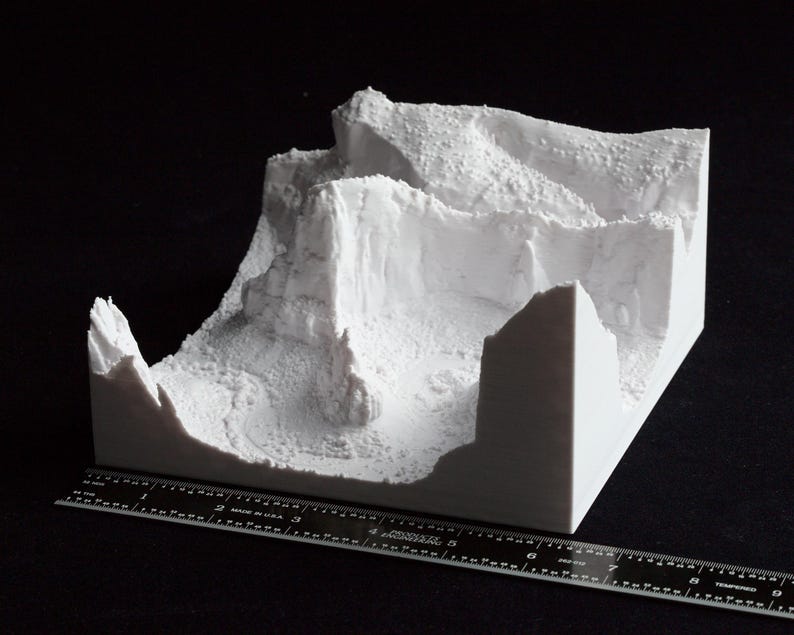 May include: A white 3D-printed model of a mountainous landscape, featuring cliffs, valleys, and textured surfaces. A ruler is placed next to the model for scale. The model is on a black surface.