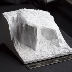 May include: A white 3D printed model of a rocky landscape with a large, jagged cliff face. The model is textured to resemble rock and is sitting on a black surface.
