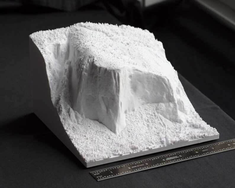 May include: A white 3D printed model of a rocky landscape with a large, jagged cliff face. The model is textured to resemble rock and is sitting on a black surface.