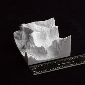 May include: A white 3D-printed model of a mountainous landscape. The model has a textured surface and is placed on a black surface. A ruler with inch measurements is next to the model, showing its size.