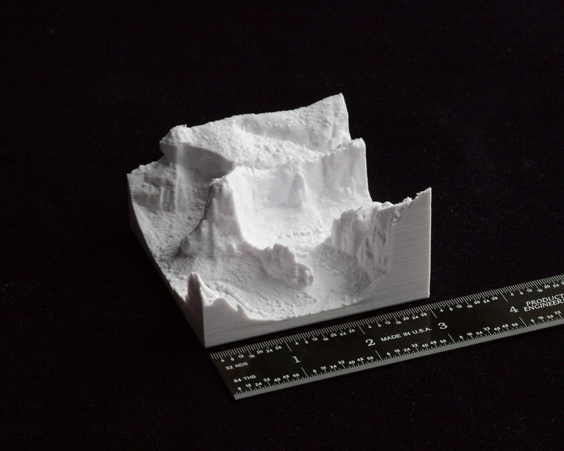 May include: A white 3D-printed model of a mountainous landscape. The model has a textured surface and is placed on a black surface. A ruler with inch measurements is next to the model, showing its size.