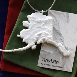 May include: A white 3D printed model of the state of Alaska, hanging from a silver chain. The model is on a green velvet surface with a white card that reads "TinyMtn 3D Printed Landscapes".
