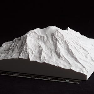 May include: A white, 3D-printed model of a mountain range. The model has detailed textures and a flat base. A ruler is placed in front of the model, with the mountain range against a black background.