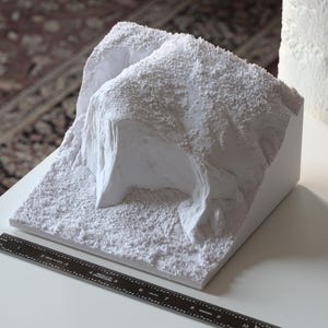 May include: A white, textured, 3D printed model of a rocky landscape. The model is on a white surface with a black ruler.
