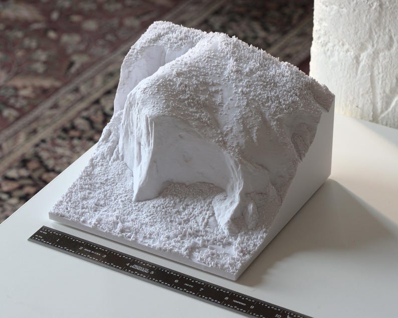 May include: A white, textured, 3D printed model of a rocky landscape. The model is on a white surface with a black ruler.