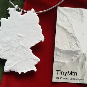 Pode incluir: Um pingente branco impresso em 3D com a forma da Alemanha, pendurado em uma corrente prateada. O pingente está sobre um fundo de veludo vermelho. Um cartão branco com o texto "TinyMtn 3D Printed Landscapes" está no fundo.