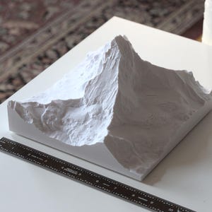 Puede incluir: Modelo de pico de monta&ntilde;a de yeso blanco, con una superficie texturizada, sobre una base blanca. El modelo mide aproximadamente 25 cm de ancho.