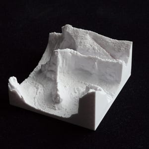 May include: A small, white, 3D-printed model of a mountainous landscape. The model features layered terrain with cliffs and valleys, showcasing a detailed, textured surface. The object is set against a black background.