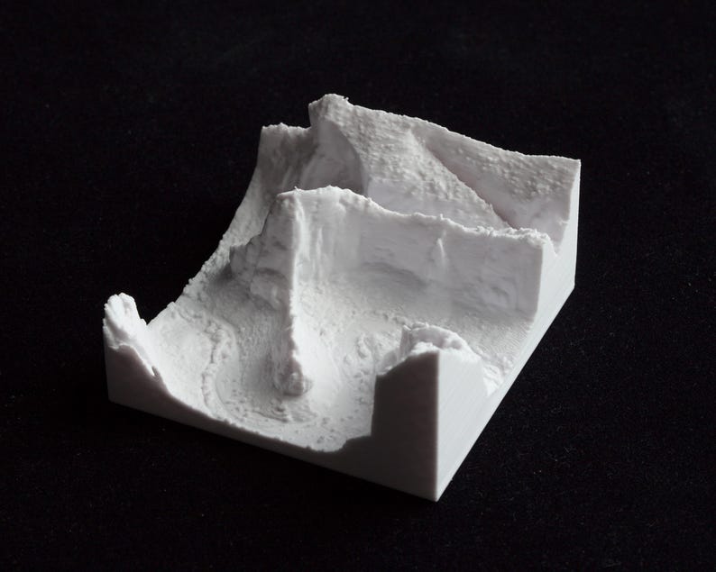 May include: A small, white, 3D-printed model of a mountainous landscape. The model features layered terrain with cliffs and valleys, showcasing a detailed, textured surface. The object is set against a black background.