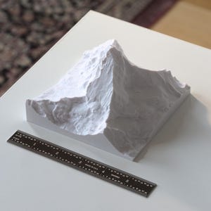 Puede incluir: Un modelo 3D impreso en blanco de una cordillera de monta&ntilde;as sobre una superficie blanca con una regla negra.