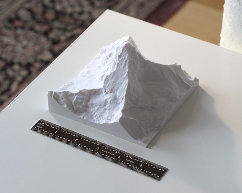 Puede incluir: Un modelo 3D impreso en blanco de una cordillera de monta&ntilde;as sobre una superficie blanca con una regla negra.