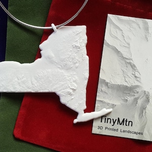 Puede incluir: Un colgante blanco impreso en 3D con la forma del estado de Nueva York, con una superficie topográfica detallada. El colgante cuelga de una cadena plateada. También se ve una tarjeta de visita con una imagen de paisaje montañoso y el texto "TinyMtn 3D Printed Landscapes".