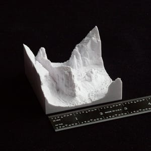 May include: A white 3D-printed model of a mountain range. The model is on a black surface, with a ruler next to it for scale. The model appears to be approximately 3 inches long.
