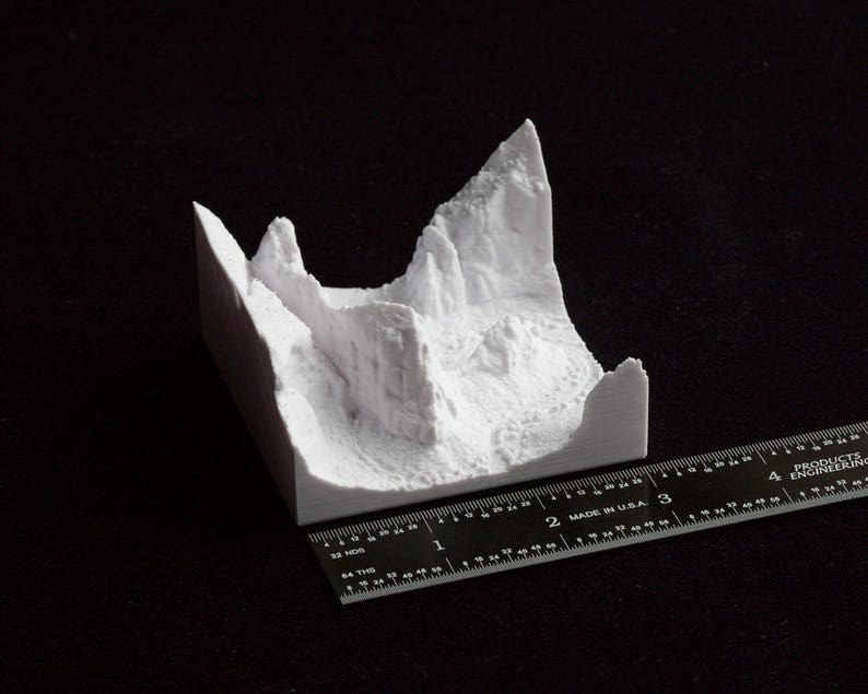 May include: A white 3D-printed model of a mountain range. The model is on a black surface, with a ruler next to it for scale. The model appears to be approximately 3 inches long.