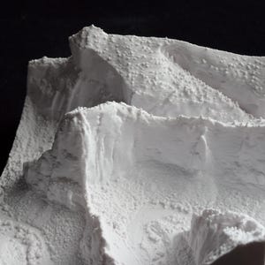 May include: A detailed, white miniature landscape model with textured surfaces, resembling a mountainous terrain. The model features layered elevations and rough, rocky formations, creating a realistic depiction of a natural environment. The model is set against a dark background.