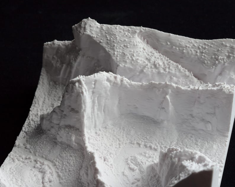 May include: A detailed, white miniature landscape model with textured surfaces, resembling a mountainous terrain. The model features layered elevations and rough, rocky formations, creating a realistic depiction of a natural environment. The model is set against a dark background.