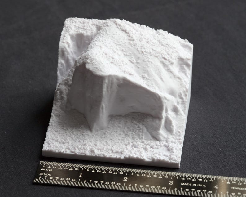 May include: White 3D printed model of a rocky cliff face. The model is textured to resemble a natural rock formation.