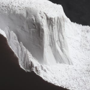 May include: A close-up of a white, textured, 3D printed model of a mountain range. The model is made of a fine, powdery material and has a rough, uneven surface.