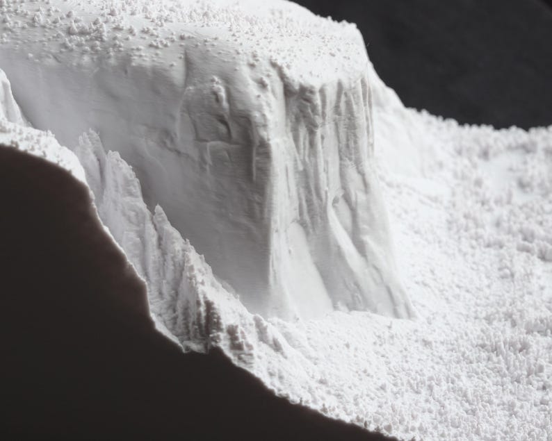 May include: A close-up of a white, textured, 3D printed model of a mountain range. The model is made of a fine, powdery material and has a rough, uneven surface.