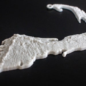 May include: A white 3D printed outline of Cape Cod, a popular vacation destination in Massachusetts. The outline is detailed and textured, resembling the shape of the peninsula.