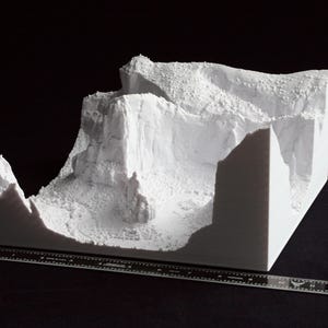 May include: A white 3D-printed model of a mountainous landscape, featuring cliffs and textured terrain. A ruler is placed in front of the model for scale. The model is set against a black background.