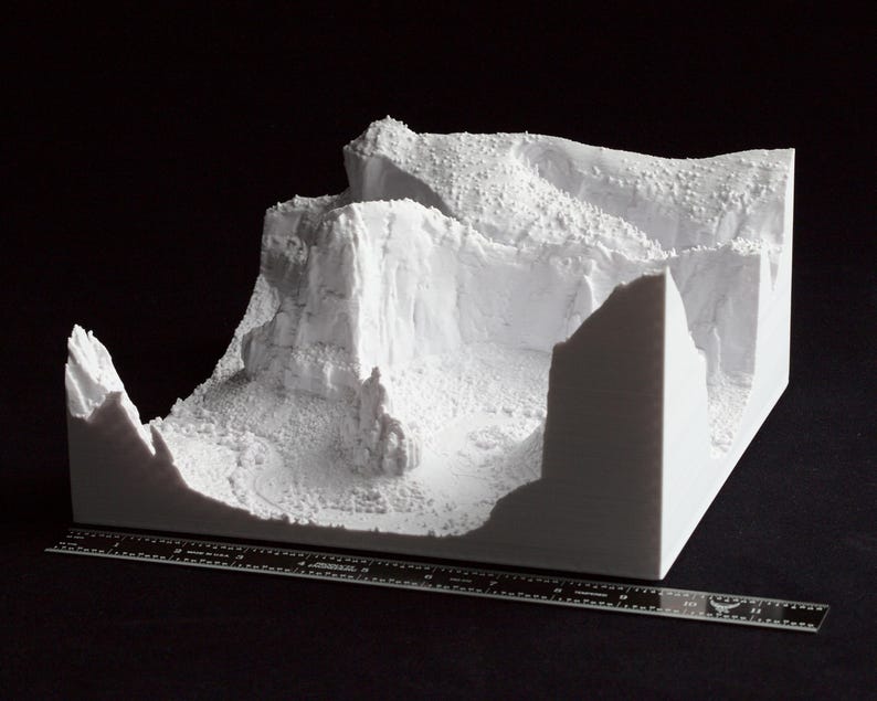 May include: A white 3D-printed model of a mountainous landscape, featuring cliffs and textured terrain. A ruler is placed in front of the model for scale. The model is set against a black background.