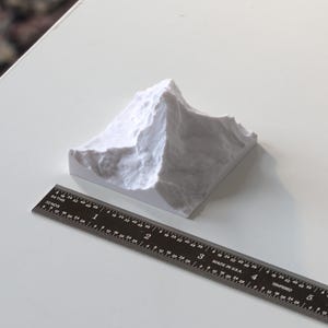 Puede incluir: Un modelo blanco de una cordillera de monta&ntilde;as impreso en 3D. El modelo mide aproximadamente 5 cm de alto y 7,5 cm de ancho. Una regla est&aacute; en primer plano para la escala.