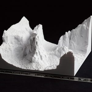 May include: A white 3D-printed model of a mountainous landscape, featuring peaks, valleys, and textured surfaces. A ruler is placed in front of the model for scale. The model is set against a black background.