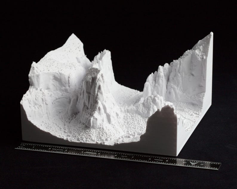 May include: A white 3D-printed model of a mountainous landscape, featuring peaks, valleys, and textured surfaces. A ruler is placed in front of the model for scale. The model is set against a black background.
