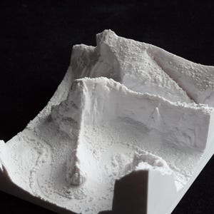 May include: A white, 3D-printed topographical model of a mountainous landscape. The model features detailed textures and contours, with peaks, valleys, and a winding path. The overall design is a square shape, and the model is set against a black background.