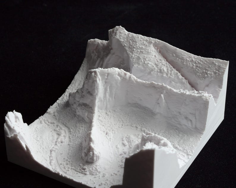 May include: A white, 3D-printed topographical model of a mountainous landscape. The model features detailed textures and contours, with peaks, valleys, and a winding path. The overall design is a square shape, and the model is set against a black background.