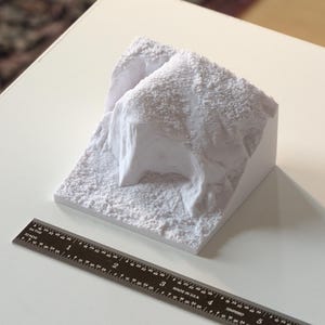May include: A white 3D printed model of a rocky terrain. The model is about 4 inches wide and 3 inches tall. It is made of a smooth, white plastic.