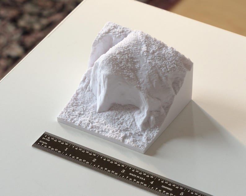May include: A white 3D printed model of a rocky terrain. The model is about 4 inches wide and 3 inches tall. It is made of a smooth, white plastic.