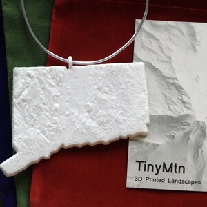 May include: White 3D printed model of the state of Connecticut hanging from a silver cord. The model is textured to resemble mountains. The background is a red fabric with a white card that reads "TinyMtn 3D Printed Landscapes".