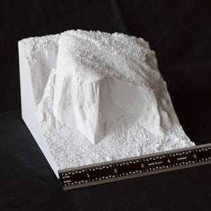 May include: White plaster model of a rocky cliff face with a textured surface. A ruler is placed at the bottom of the model for scale.