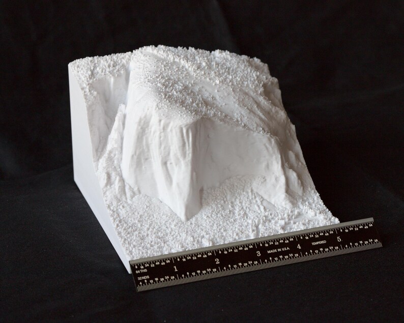 May include: White plaster model of a rocky cliff face with a textured surface. A ruler is placed at the bottom of the model for scale.