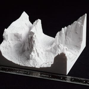 May include: A white 3D-printed model of a mountainous landscape, featuring peaks, valleys, and cliffs. The model is placed on a black surface with a ruler for scale. The terrain is detailed with textured surfaces.