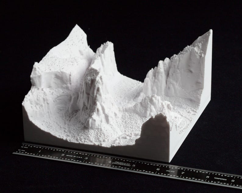 May include: A white 3D-printed model of a mountainous landscape, featuring peaks, valleys, and cliffs. The model is placed on a black surface with a ruler for scale. The terrain is detailed with textured surfaces.
