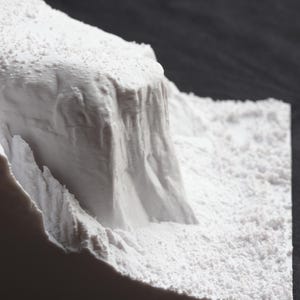 May include: A close-up of a white, textured, 3D printed model of a mountain range. The model is made of a smooth, white material and has a rough, textured surface.